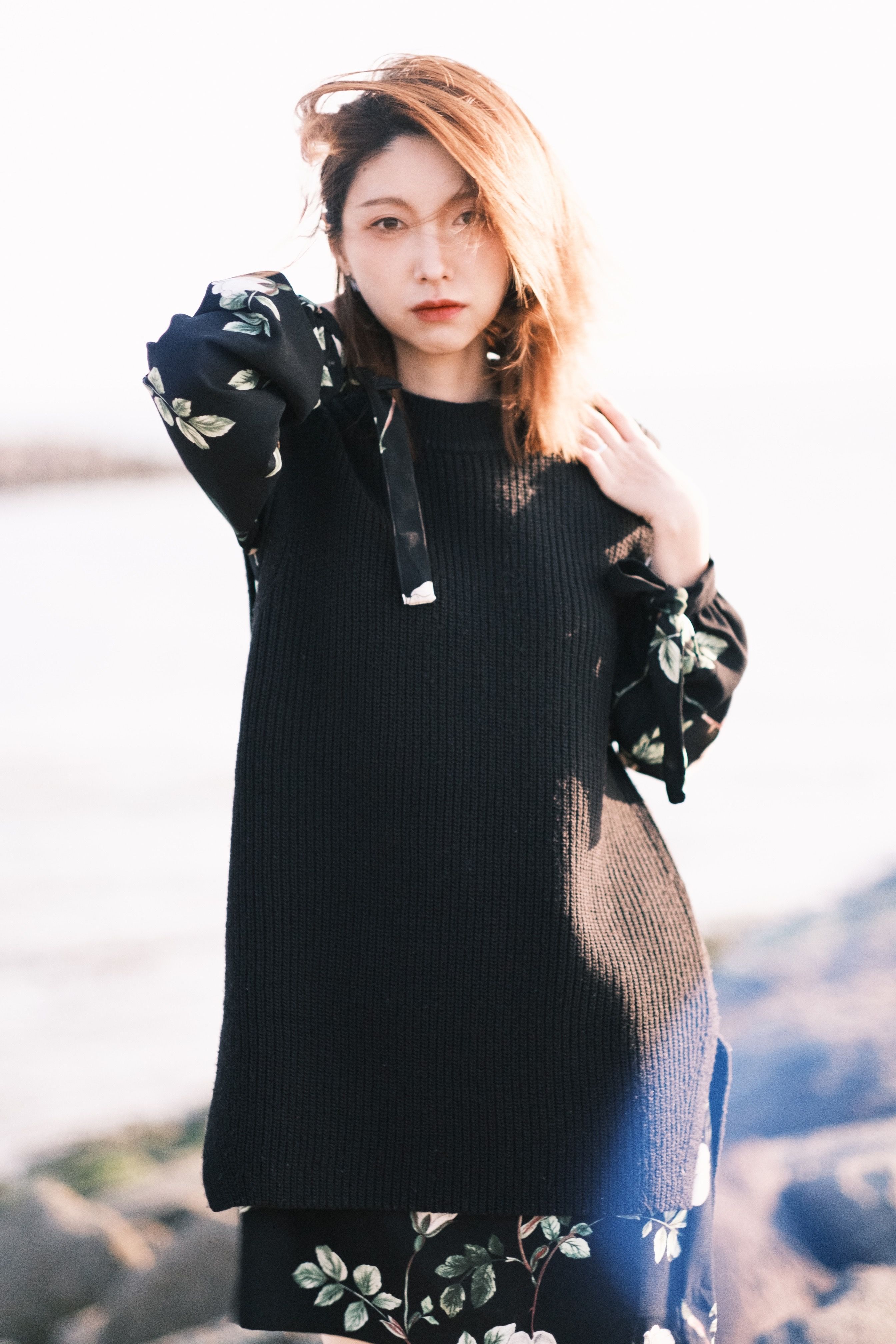 Calm young Asian female in stylish outfit standing on rocky beach near sea and looking at camera