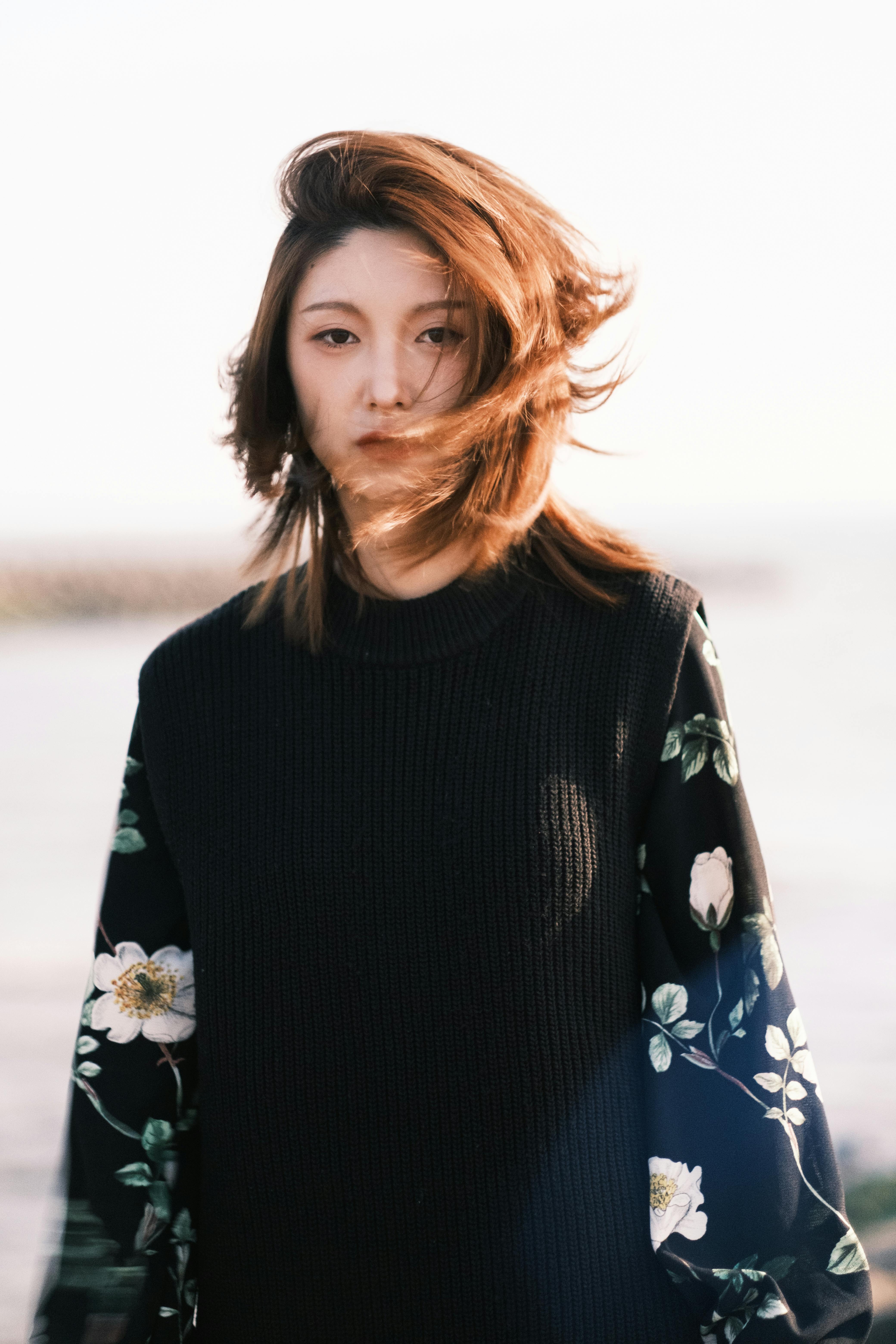 Dreamy young ethnic female tourist with brown hair in elegant outfit relaxing on seashore on windy day and looking at camera