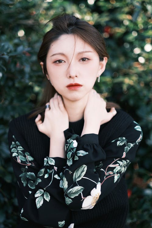 Thoughtful young Asian lady with brown hair touching neck and looking at camera while resting in lush green garden on sunny day