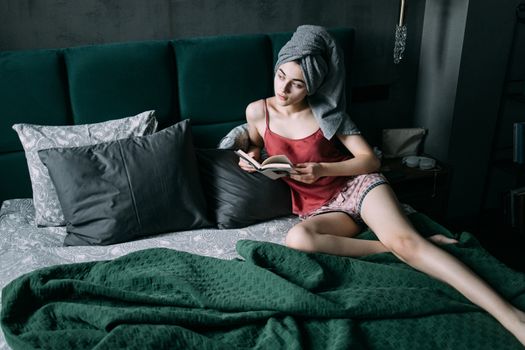 Woman in bedroom with towel on head, enjoying a book on bed. Comfortable and serene indoor setting.