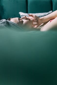 Woman lying in bed with a towel turban, applying facial skincare with eyes closed.