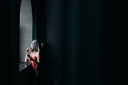 Young woman enjoying self-care with a clay mask, sipping tea by the window.