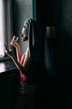 Woman with face mask and towel enjoying skincare routine by window, holding a smartphone and glass.