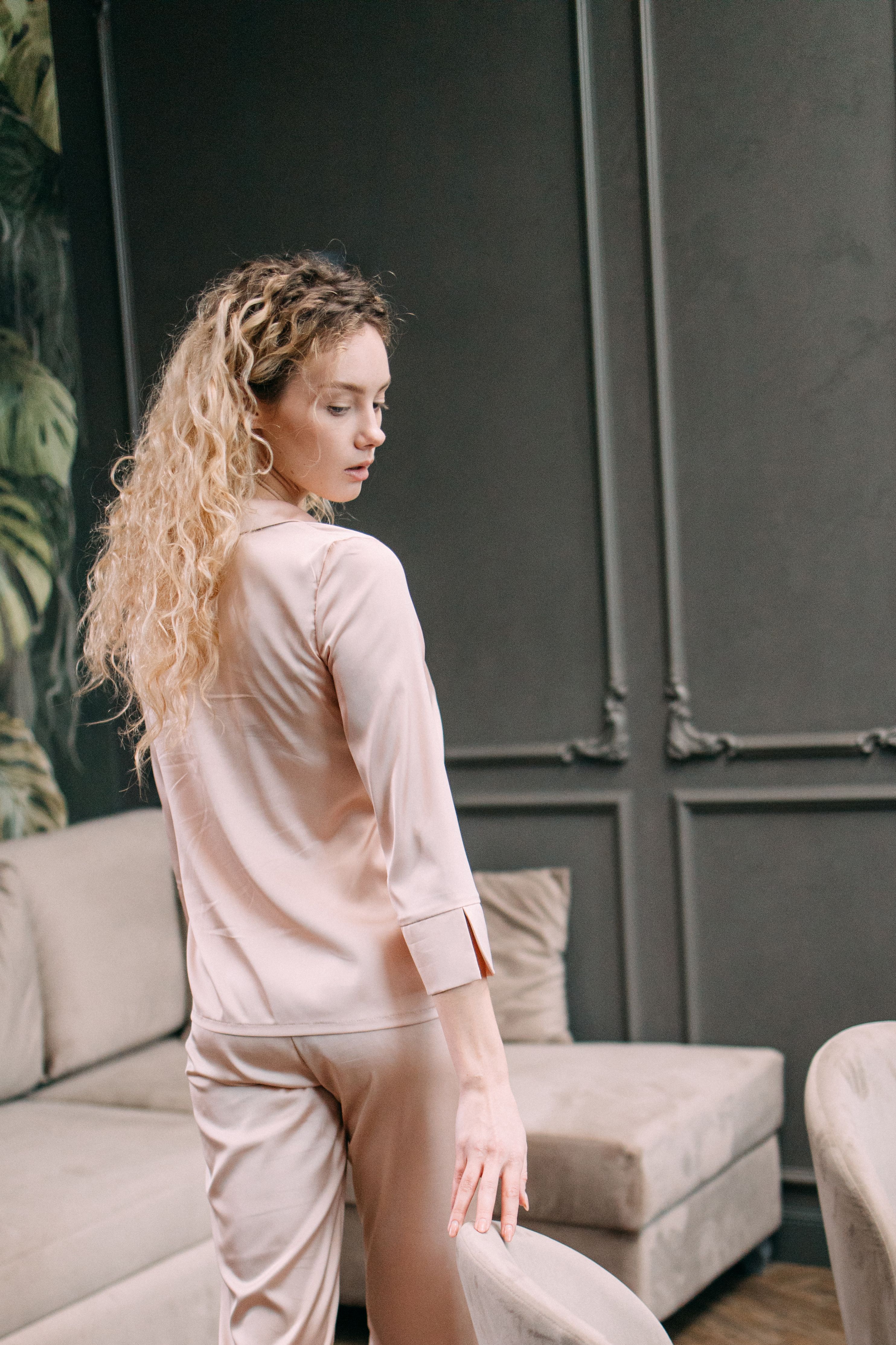 Mulher estilosa com cabelo loiro encaracolado em pijama elegante, posando de forma sofisticada no interior, criando uma atmosfera graciosa e sofisticada.
