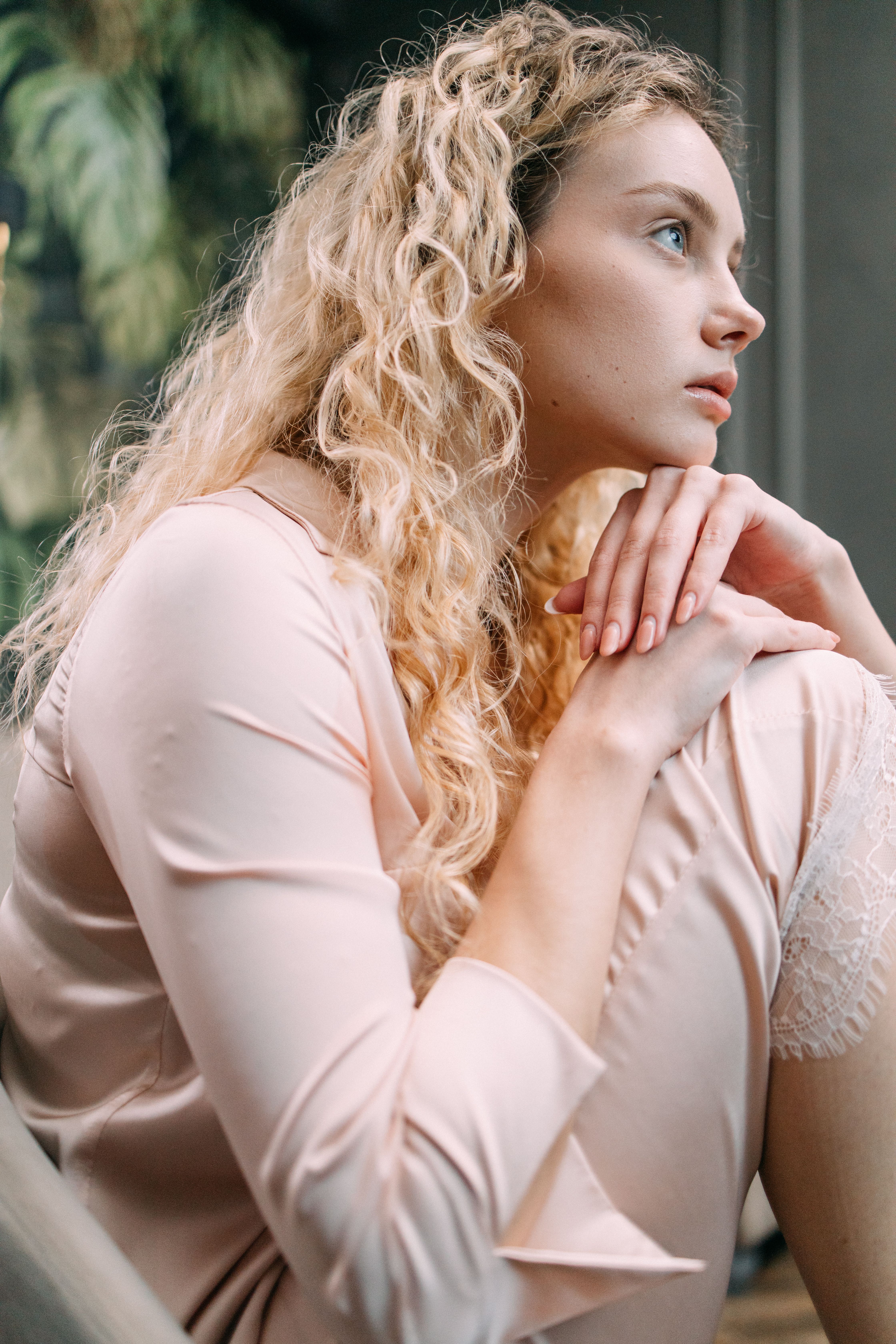 A thoughtful young woman in loungewear sits indoors, gazing out with a serene expression.