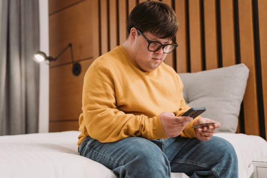 Man with Down syndrome in a yellow sweater sits on a bed using a mobile phone, indoors.