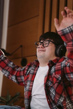 A happy man with headphones and glasses joyfully raising his hands indoors.