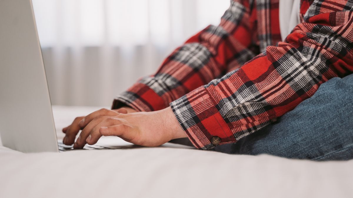 Close-up of hands typing on a laptop, wearing a red plaid shirt indoors.
