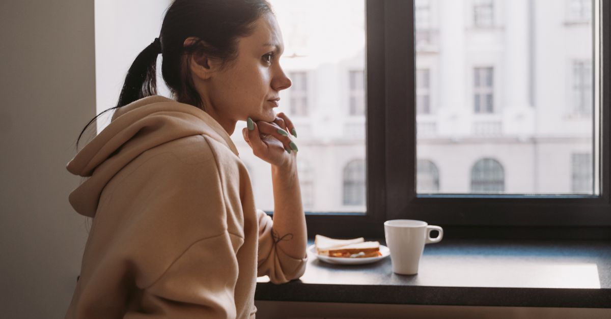Side view of a woman in a hoodie sitting by a window, contemplating while looking outside.