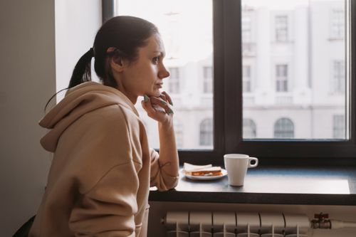 Free Side view of a woman in a hoodie sitting by a window, contemplating while looking outside. Stock Photo