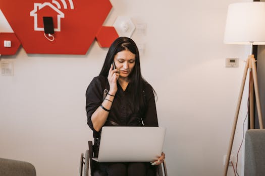 Woman in a wheelchair multitasking with a laptop and phone in a modern home setting.