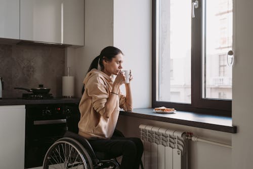 Free A Woman Sitting on Her Wheelchair while Drinking Coffee Near the Glass Window Stock Photo
