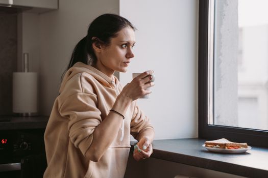 Woman in a hoodie enjoying a hot drink by the window with a plate of food nearby.