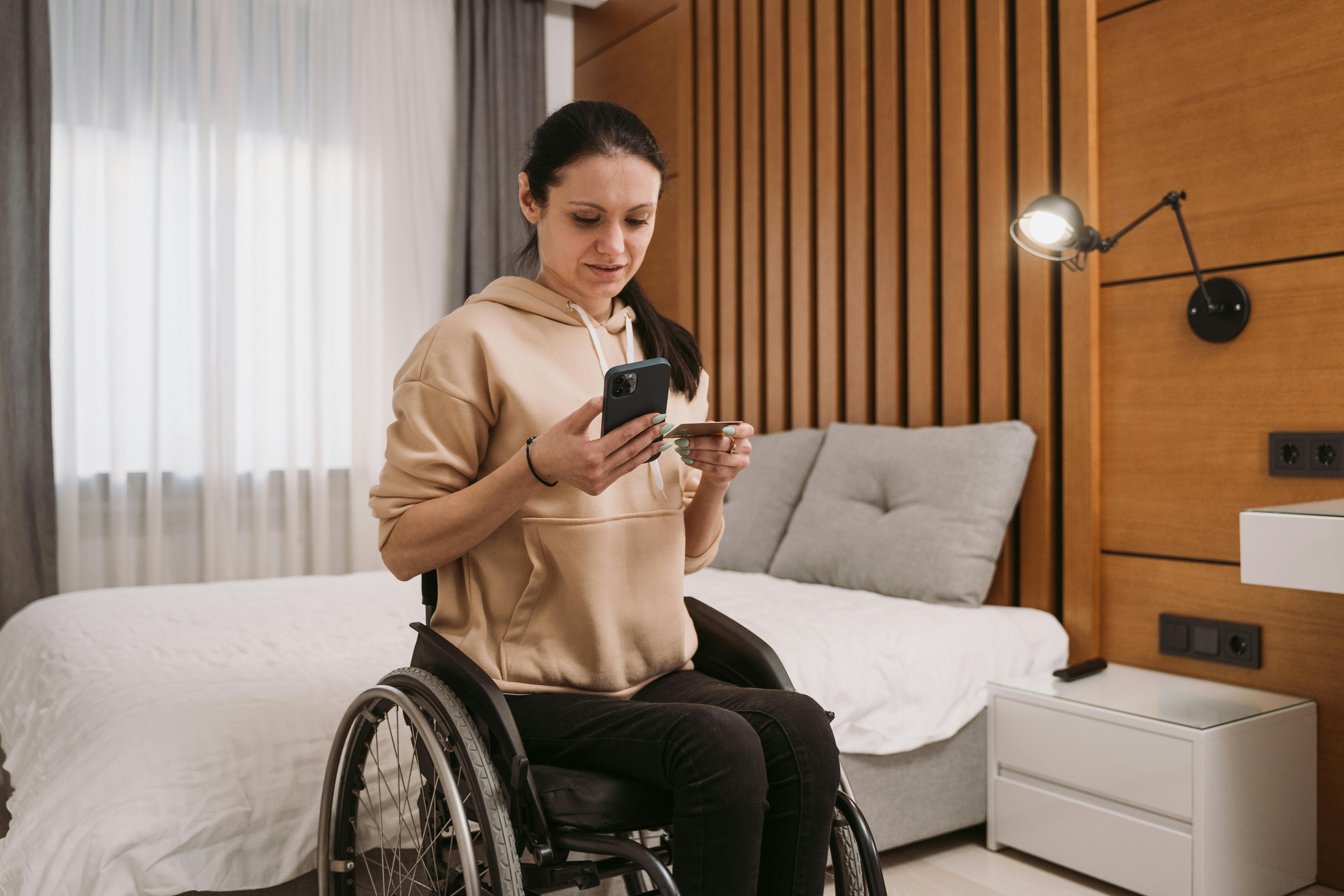 Woman in a wheelchair using a smartphone and credit card in a modern bedroom.