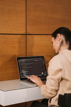 Young woman typing on a laptop while coding in a modern indoor setting with a wooden wall background.