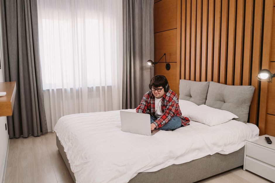 A young man with headphones engaged on a laptop while sitting on a bed in a modern bedroom. - How to Stop Fitted Sheets From Popping Off: 4 Elastic Corner Fasteners Tested