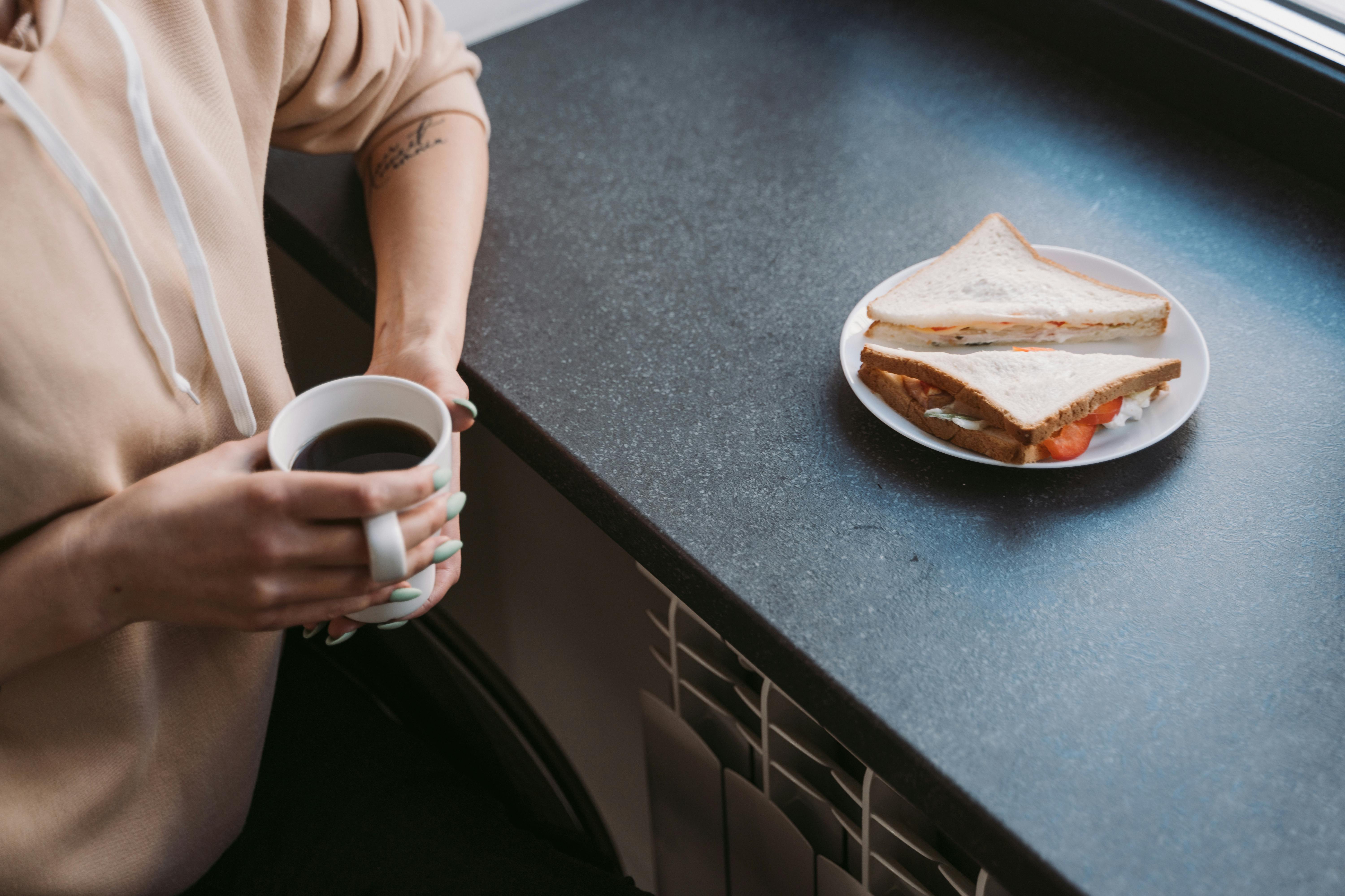 Person in hoodie enjoying coffee and sandwich by the window.