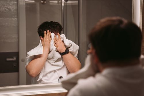 Free A man standing in front of a bathroom mirror, wiping his face with a towel, emphasizing personal hygiene. Stock Photo