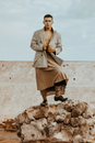 Full body of confident Asian male wearing authentic skirt and blazer looking at camera while standing on heap of rocks in countryside