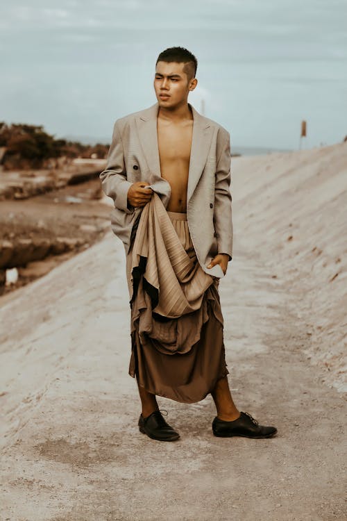 Full body of confident Asian male in blazer and traditional skirt looking away while standing in suburb area in countryside