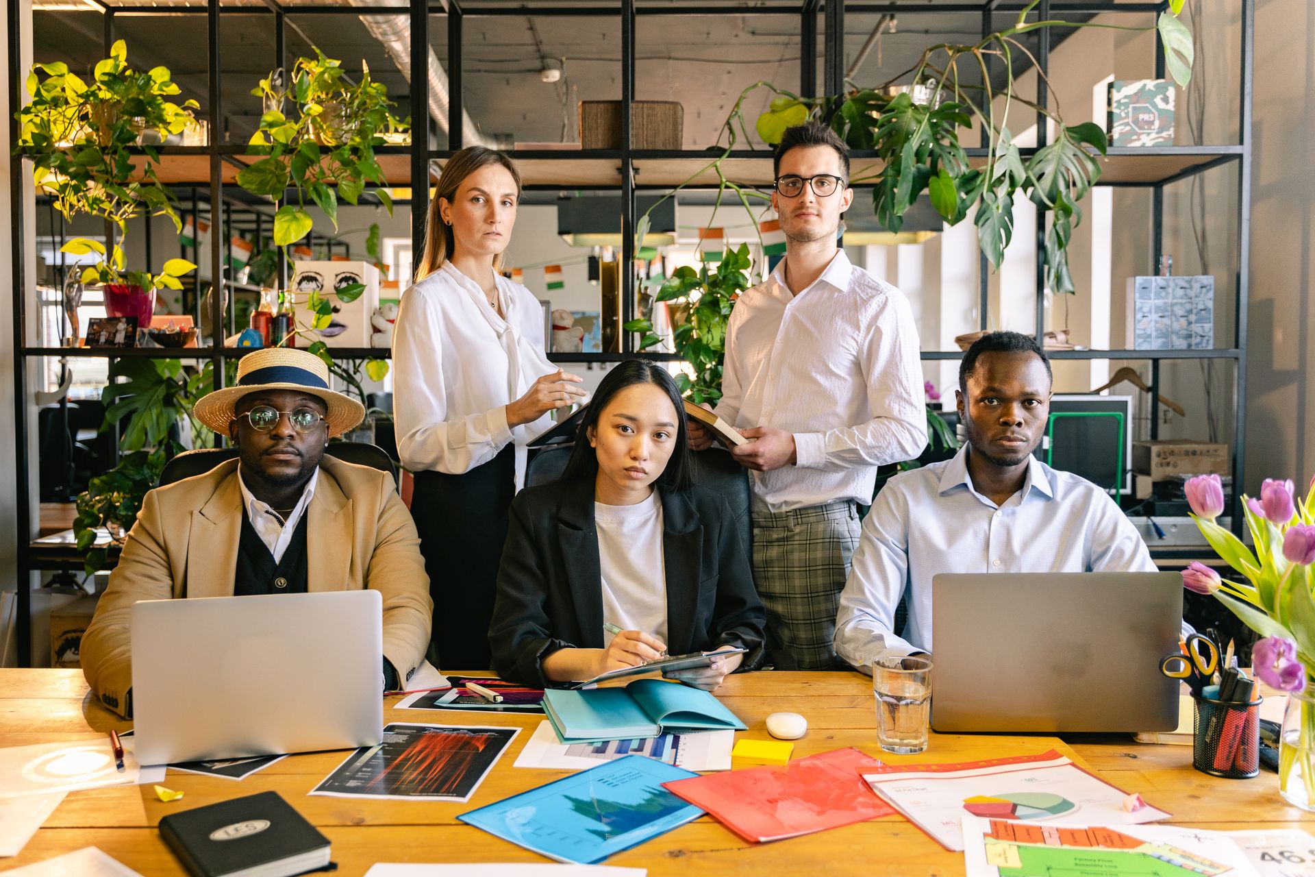 A diverse group of business professionals working together in a modern, plant-filled office space with laptops and documents.