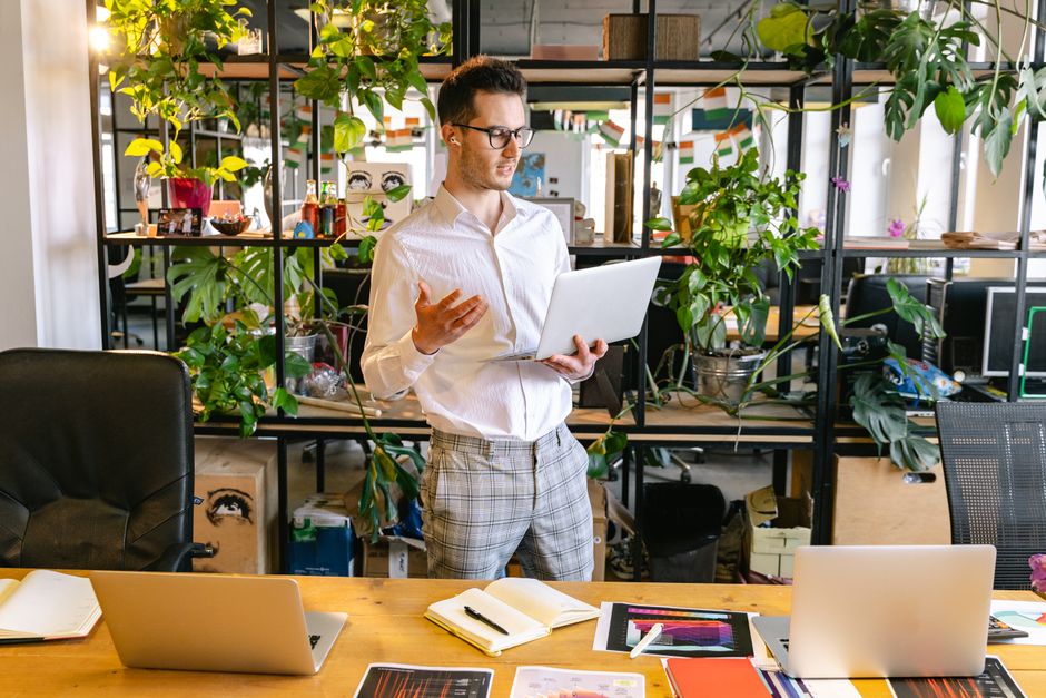 Professional man having a video call in a green indoor office space with multiple laptops and potted plants.