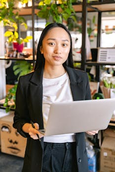 Confident woman in a black blazer using a laptop in a modern office setting with greenery.