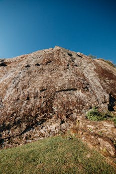 Rocky mountain with rough surface on grassy terrain against clear cloudless blue sky in suburb area on sunny summer day