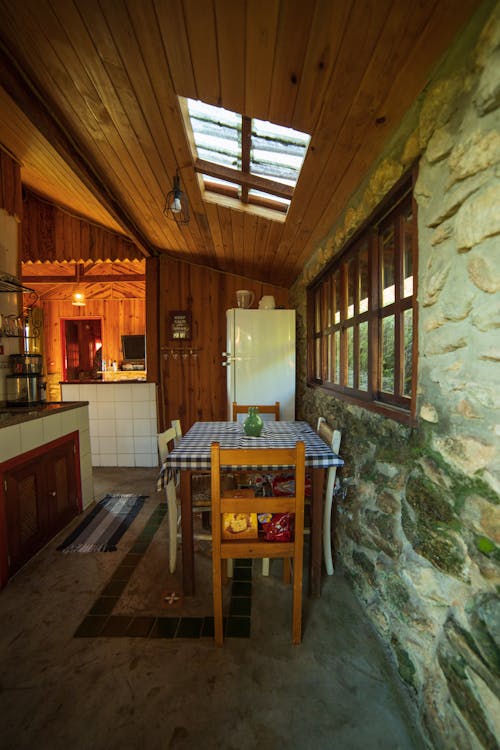 Chairs at table with tablecloth placed near fridge at wall with window decorated with stone in rural house with wooden ceiling