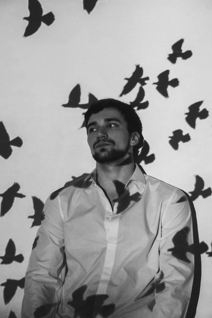 Black and white of dreamy young male in white shirt leaning on wall with shadows of flying birds while looking away
