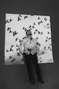 Black and white full body of male in white shirt standing in dark studio near wall with shadows of birds flying under sky