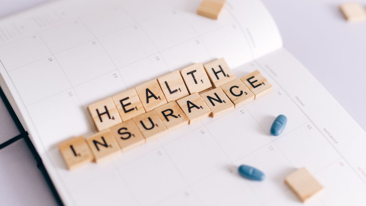 Close-up of health insurance spelled out with Scrabble tiles on a planner, alongside medication pills.
