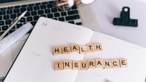 Close-up of Scrabble tiles spelling 'Health Insurance' on a planner with a laptop.