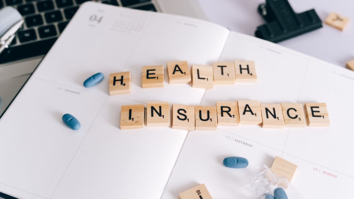 Scrabble tiles spelling 'Health Insurance' on planner with pills and laptop, symbolizing healthcare planning.