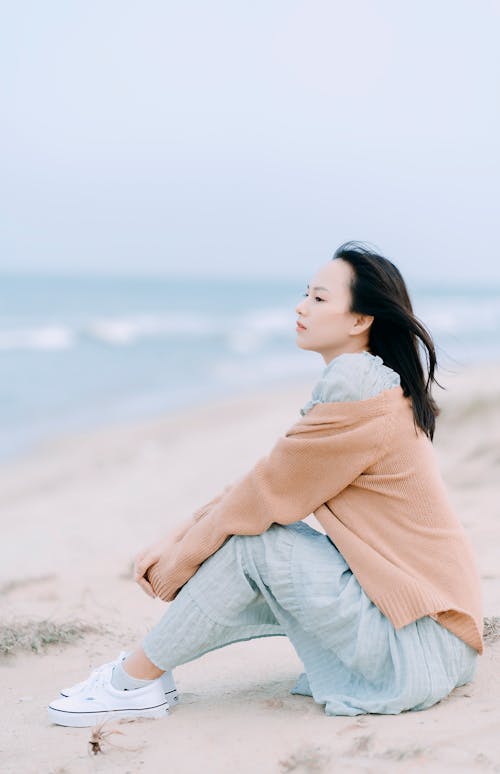 Full body side view of pensive Asian female sitting on sandy coast near waving sea in nature in windy weather