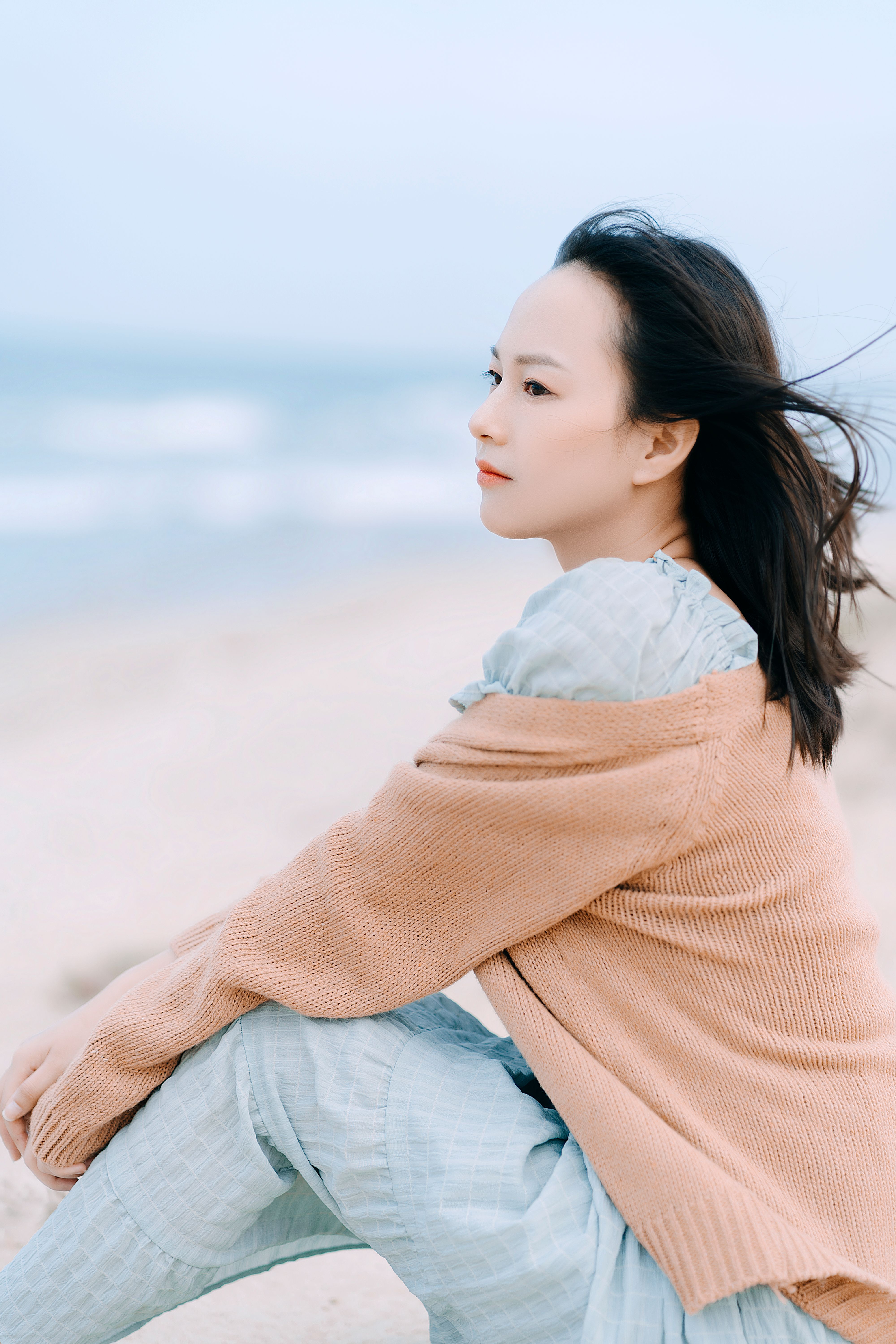 Side view of pensive Asian female traveler in casual wear sitting on sandy shore near waving sea in nature on blurred background