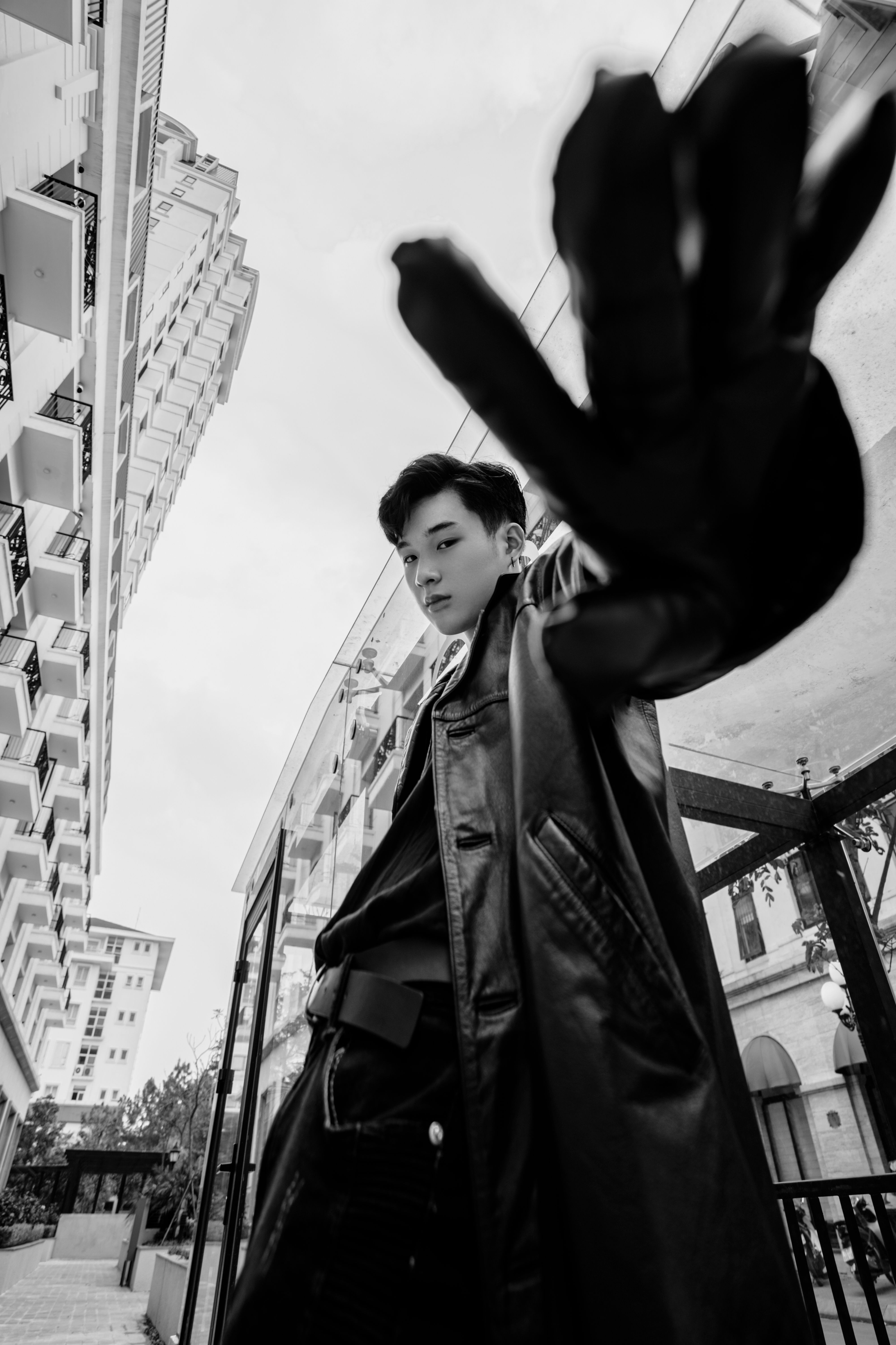 Black and white from below of confident Asian male in trendy outfit reaching hand and looking at camera while standing on street