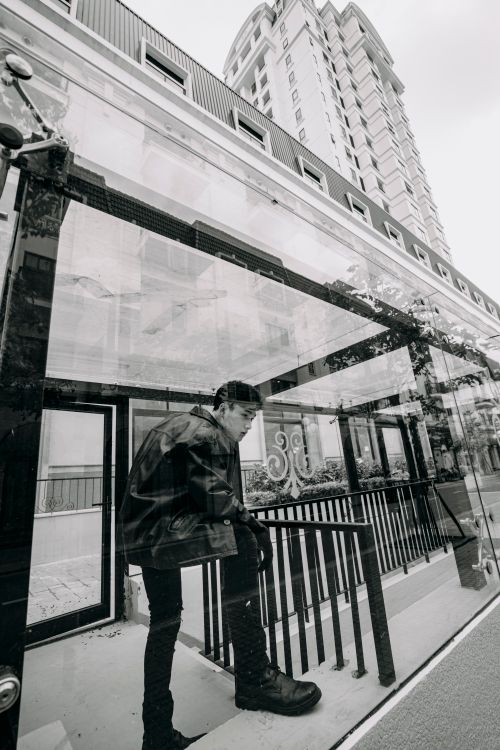 Free Black and white side view of Asian male in stylish outfit standing on bus stop in city with tall building on street Stock Photo