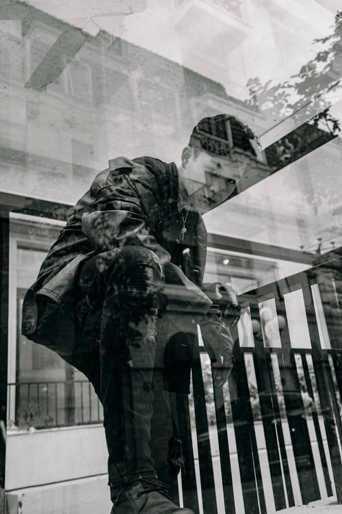 Free Black and white through window of Asian male in trendy outfit and leather jacket looking at camera while standing on bus stop in city Stock Photo