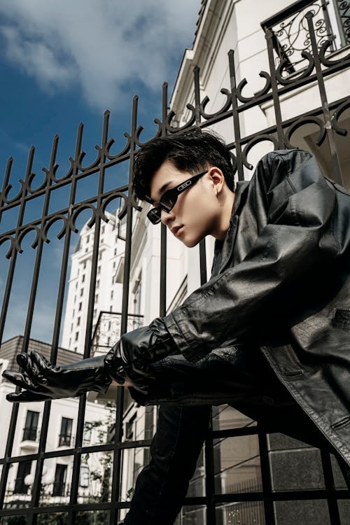 Low angle side view of trendy ethnic man in leather jacket putting on glove against urban houses