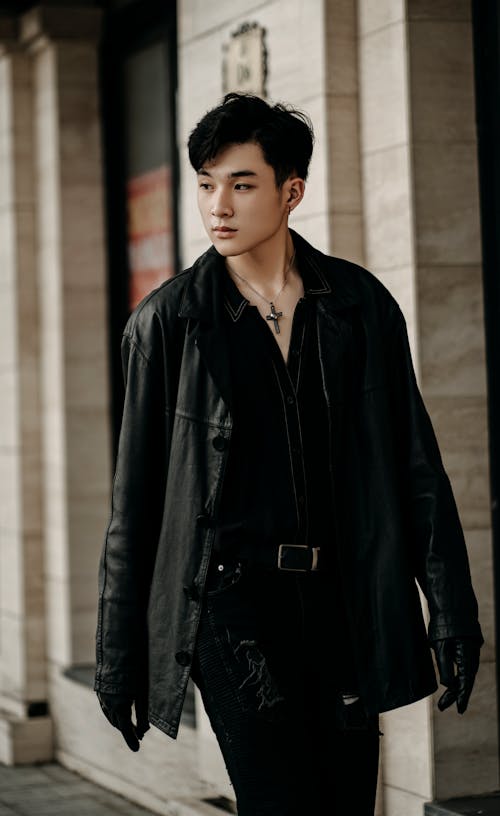 Confident young Asian male wearing black outfit and leather jacket strolling on city street and looking away