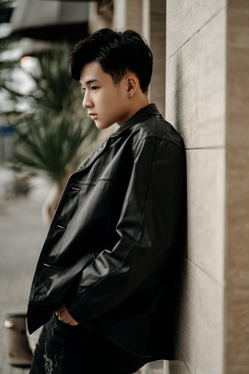 Side view contemplative young Asian male in black leather jacket standing with hands in pockets on modern city street
