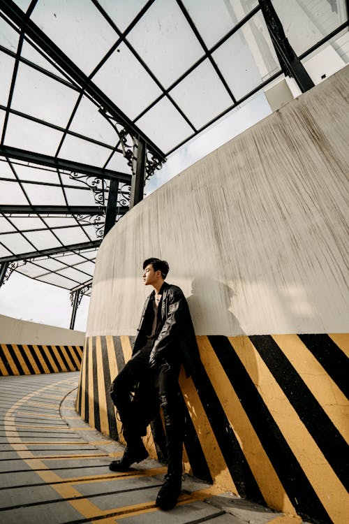Full body unemotional trendy Asian male in black outfit standing in roofed passage with glass ceiling and looking away