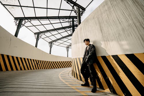 Full length self assured young Asian male in black outfit and leather jacket standing in parking passage with glass ceiling and looking away