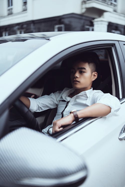 Free Confident successful Asian male in classy white shirt sitting on modern white car drivers seat and looking away Stock Photo