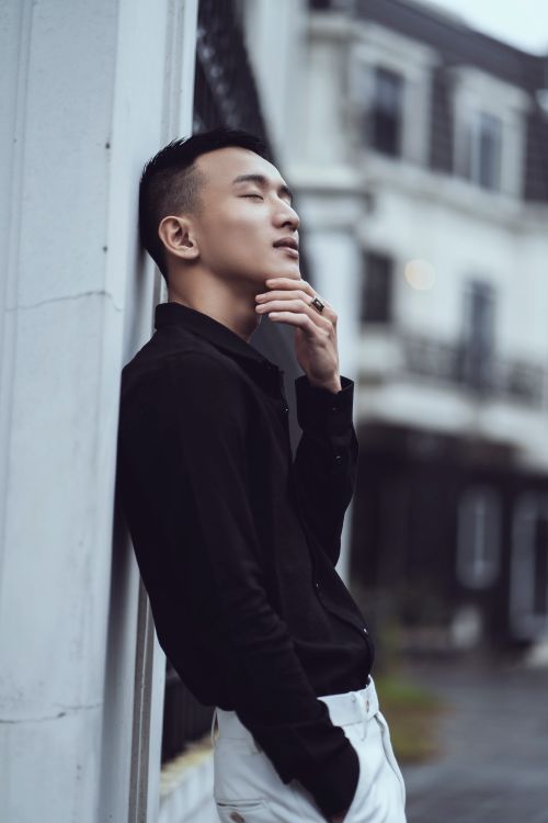 Free Side view positive young Asian male in trendy clothes touching chin and standing with eyes closed and hand in pocket on street Stock Photo