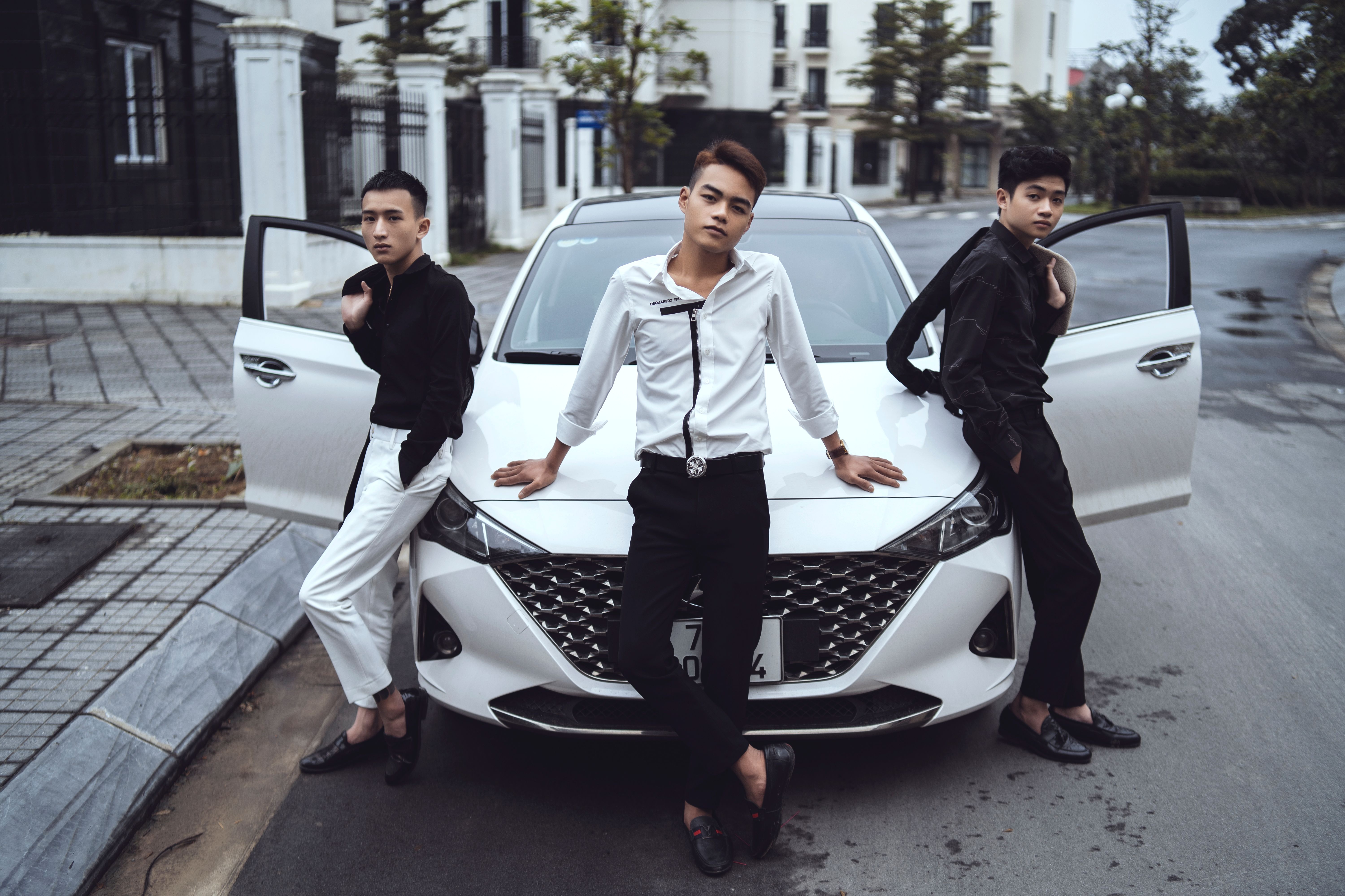 Full body of confident stylish Asian male friends in classy clothes leaning on modern car parked on city street and looking at camera