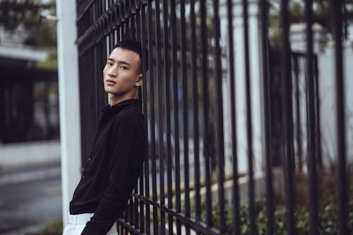 Serious young Asian guy leaning on fence on street