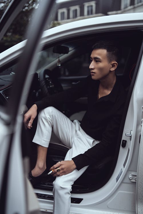 Side view of self confident young ethnic man in fashionable outfit with cigarette in hand sitting in modern car with opened door and looking away