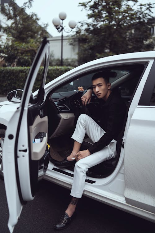 Free Full body of young relaxed Asian man in stylish clothes leaning on steering wheel and looking at camera while sitting in contemporary car with opened doors Stock Photo